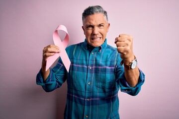 Middle age handsome grey-haired man holding pink cancer ribbon over isolated background annoyed and frustrated shouting with anger, crazy and yelling with raised hand, anger concept