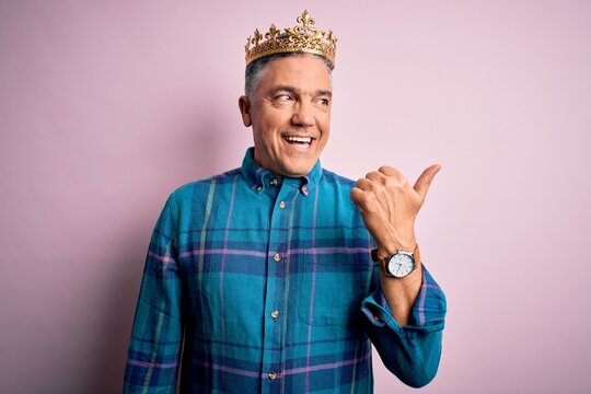 Middle age handsome grey-haired man wearing golden crown of king over pink background smiling with happy face looking and pointing to the side with thumb up.