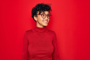 Fototapeta na wymiar Young beautiful african american afro woman wearing turtleneck sweater and glasses looking away to side with smile on face, natural expression. Laughing confident.
