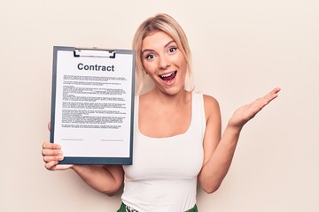 Beautiful blonde woman holding clipboard with contract document paper over white background celebrating achievement with happy smile and winner expression with raised hand