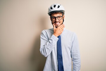 Young businessman wearing glasses and bike helmet standing over isolated white bakground looking confident at the camera smiling with crossed arms and hand raised on chin. Thinking positive.
