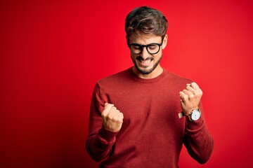 Young handsome man with beard wearing glasses and sweater standing over red background celebrating...