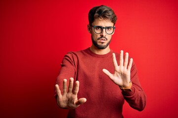 Young handsome man with beard wearing glasses and sweater standing over red background Moving away hands palms showing refusal and denial with afraid and disgusting expression. Stop and forbidden.