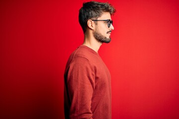 Fototapeta na wymiar Young handsome man with beard wearing glasses and sweater standing over red background looking to side, relax profile pose with natural face and confident smile.