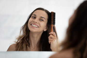 Head shot close up happy young woman brushing long dark hair after shower, happy beautiful girl with toothy smile looking in mirror, standing in bathroom, doing hairstyle, morning routine and beauty