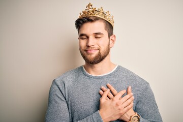 Young blond man with beard and blue eyes wearing golden crown of king smiling with hands on chest...