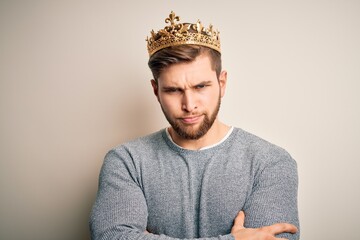 Young blond man with beard and blue eyes wearing golden crown of king skeptic and nervous, disapproving expression on face with crossed arms. Negative person.