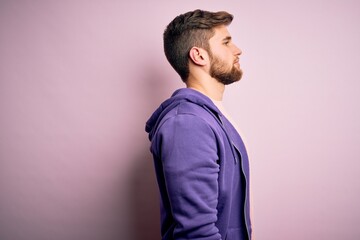 Young blond man with beard and blue eyes wearing purple sweatshirt over pink background looking to...