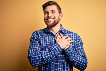 Young blond businessman with beard and blue eyes wearing shirt over yellow background smiling with hands on chest with closed eyes and grateful gesture on face. Health concept.