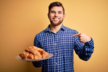 Young blond man with beard and blue eyes holding plate with french croissants to breakfast with surprise face pointing finger to himself