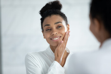 Head shot smiling African American woman touching cheek, enjoying perfect smooth face skin, looking in mirror, beautiful girl wearing white bathrobe doing facial massage in bathroom, skincare concept