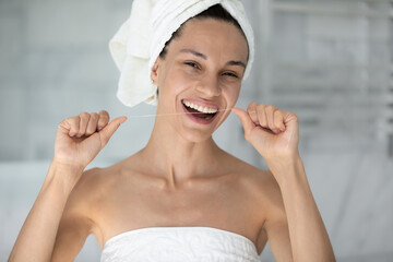 Head shot portrait beautiful woman with healthy toothy smile using mint dental floss, removing food cleaning teeth, pretty girl wearing white bath towel on head looking at camera, personal hygiene