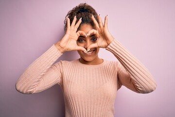 Young beautiful african american girl wearing casual sweater standing over pink background Doing heart shape with hand and fingers smiling looking through sign