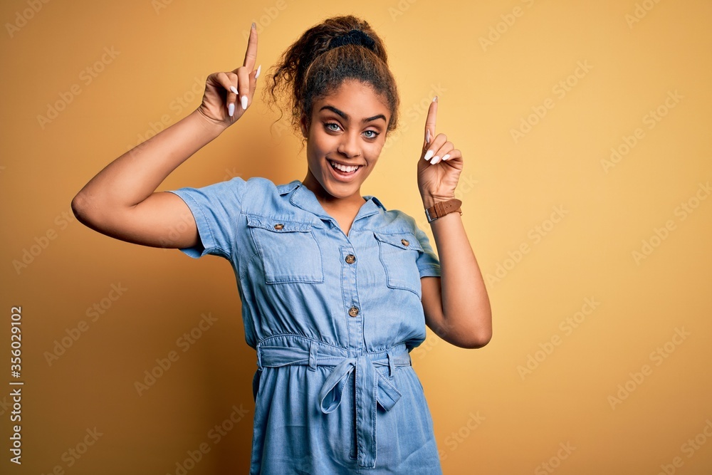 Poster Young beautiful african american girl wearing denim dress standing over yellow background smiling amazed and surprised and pointing up with fingers and raised arms.