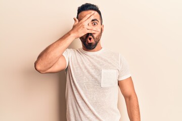 Young hispanic man wearing casual clothes peeking in shock covering face and eyes with hand, looking through fingers afraid