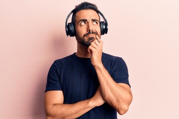 Young hispanic man listening to music using headphones serious face thinking about question with hand on chin, thoughtful about confusing idea