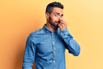 Young hispanic man wearing casual clothes smelling something stinky and disgusting, intolerable smell, holding breath with fingers on nose. bad smell - obrazy, fototapety, plakaty