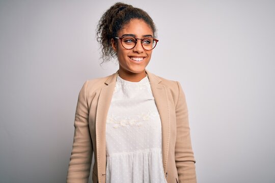 Beautiful african american businesswoman wearing jacket and glasses over white background looking away to side with smile on face, natural expression. Laughing confident.