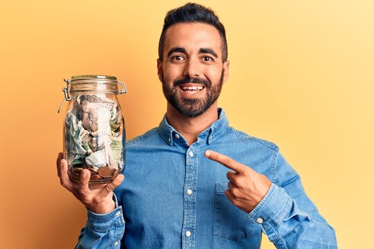 Young hispanic man holding jar with savings smiling happy pointing with hand and finger