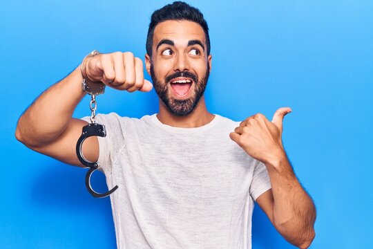 Young hispanic man holding prisoner handcuffs pointing thumb up to the side smiling happy with open mouth