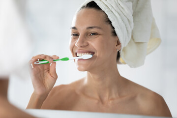 Head shot close up happy beautiful woman wearing white bath towel on head brushing teeth, looking in mirror, attractive girl with toothy smile cleaning teeth, morning routine, oral hygiene concept