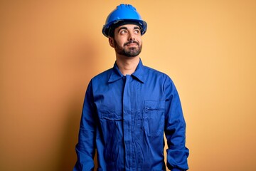 Mechanic man with beard wearing blue uniform and safety helmet over yellow background smiling looking to the side and staring away thinking.