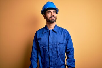 Mechanic man with beard wearing blue uniform and safety helmet over yellow background smiling...