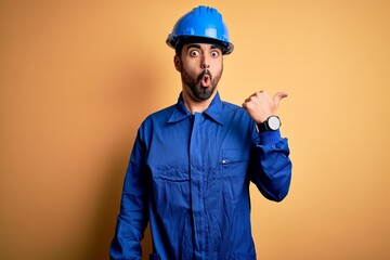 Mechanic man with beard wearing blue uniform and safety helmet over yellow background Surprised...