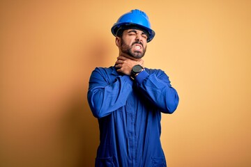 Mechanic man with beard wearing blue uniform and safety helmet over yellow background shouting suffocate because painful strangle. Health problem. Asphyxiate and suicide concept.