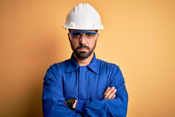 Mechanic man with beard wearing blue uniform and safety glasses over yellow background skeptic and nervous, disapproving expression on face with crossed arms. Negative person.