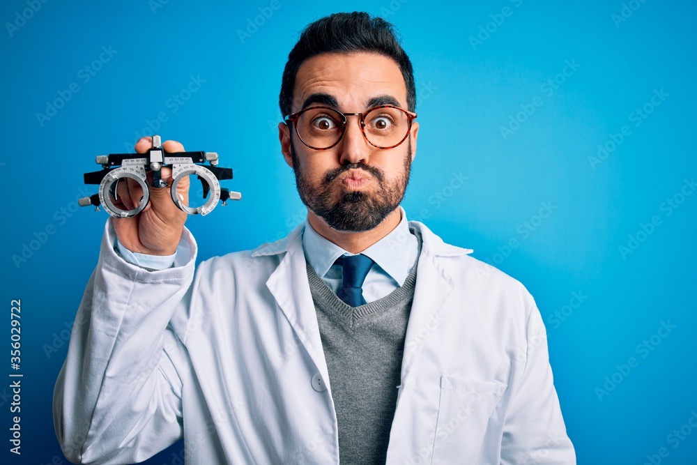 Sticker Young handsome optical man with beard holding optometry glasses over blue background puffing cheeks with funny face. Mouth inflated with air, crazy expression.