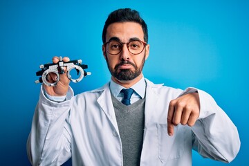 Young handsome optical man with beard holding optometry glasses over blue background Pointing down looking sad and upset, indicating direction with fingers, unhappy and depressed.