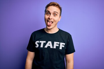 Young handsome redhead worker man wearing staff t-shirt uniform over purple background sticking tongue out happy with funny expression. Emotion concept.