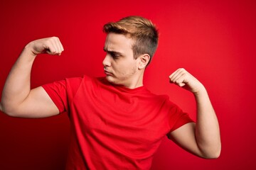 Young handsome redhead man wearing casual t-shirt over isolated red background showing arms muscles smiling proud. Fitness concept.