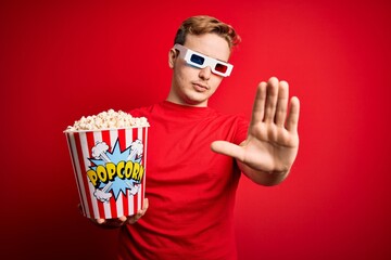 Young handsome redhead man watching 3d glasses eating popcorn snack over red background with open hand doing stop sign with serious and confident expression, defense gesture