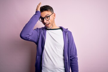 Young handsome man wearing purple sweatshirt and glasses standing over pink background surprised...