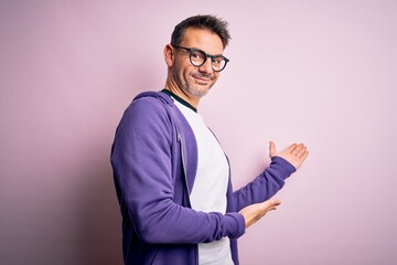 Young handsome man wearing purple sweatshirt and glasses standing over pink background Inviting to enter smiling natural with open hand