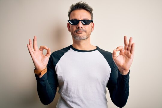 Young handsome man wearing funny thug life sunglasses meme over white background relax and smiling with eyes closed doing meditation gesture with fingers. Yoga concept.