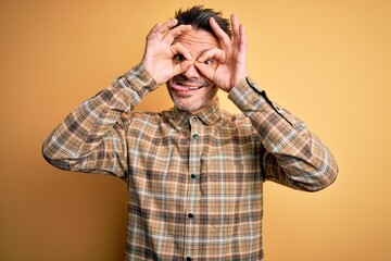 Young handsome man wearing casual shirt standing over isolated yellow background doing ok gesture like binoculars sticking tongue out, eyes looking through fingers. Crazy expression.