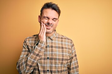 Young handsome man wearing casual shirt standing over isolated yellow background touching mouth with hand with painful expression because of toothache or dental illness on teeth. Dentist