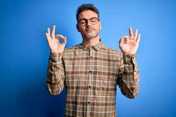Naklejka premium Young handsome man wearing casual shirt and glasses standing over blue background relax and smiling with eyes closed doing meditation gesture with fingers. Yoga concept.