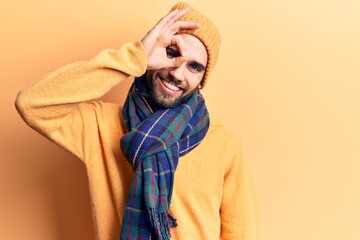 Young handsome man with beard wearing wool cap and scarf smiling happy doing ok sign with hand on eye looking through fingers