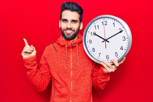 Young handsome man with beard holding big clock smiling happy pointing with hand and finger to the side