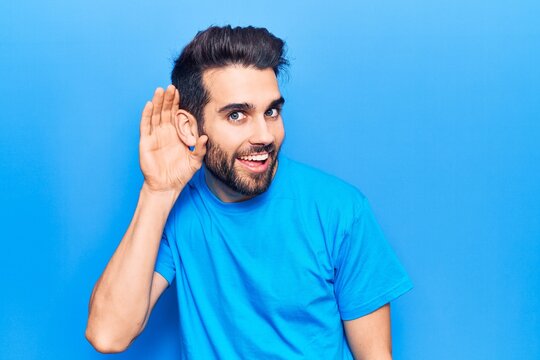 Young handsome man with beard wearing casual t-shirt smiling with hand over ear listening and hearing to rumor or gossip. deafness concept.
