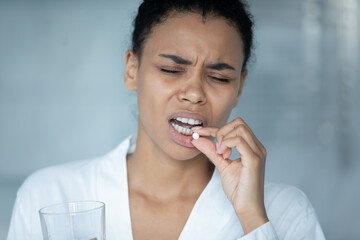Close up unhealthy exhausted African American young woman taking round white pill, holding water glass, painkiller, antidepressant or antibiotic medicine in morning, headache or depression