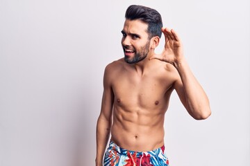 Fototapeta na wymiar Young handsome man with beard shirtless wearing swimwear smiling with hand over ear listening and hearing to rumor or gossip. deafness concept.