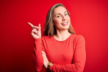 Young beautiful blonde woman wearing casual t-shirt standing over isolated red background with a big smile on face, pointing with hand and finger to the side looking at the camera.