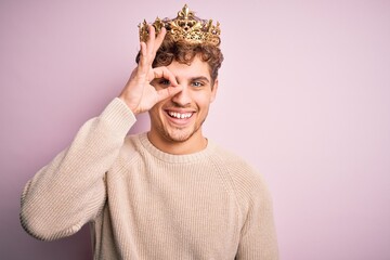 Young blond man with curly hair wearing golden crown of king over pink background doing ok gesture with hand smiling, eye looking through fingers with happy face.