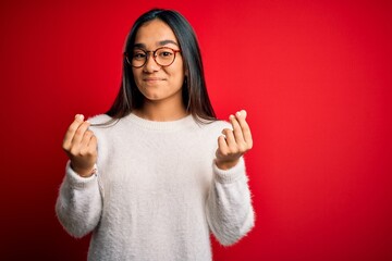 Young beautiful asian woman wearing casual sweater and glasses over red background doing money...