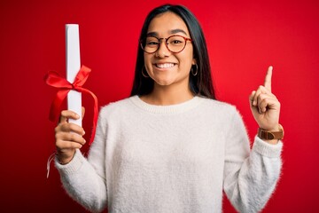 Young beautiful graduate asian woman holding university degree diploma over red background...